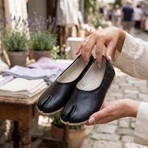 May include: A pair of black leather split-toe shoes, held by a person. The shoes have a rounded toe and a separate compartment for the big toe. The soles are a light tan color. The background shows an outdoor market setting.