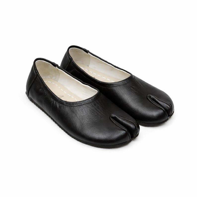 May include: A pair of black leather split-toe ballet flats. The shoes have a rounded toe and a split design for the big toe. The interior is white, and the shoes are set against a white background.