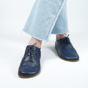 以下が含まれることがあります： レースアップクロージャーと装飾的なブローグディテールを備えたネイビーブルーの革製のオックスフォードシューズのペア。靴はライトブルーのジーンズを履いた人が履いています。
