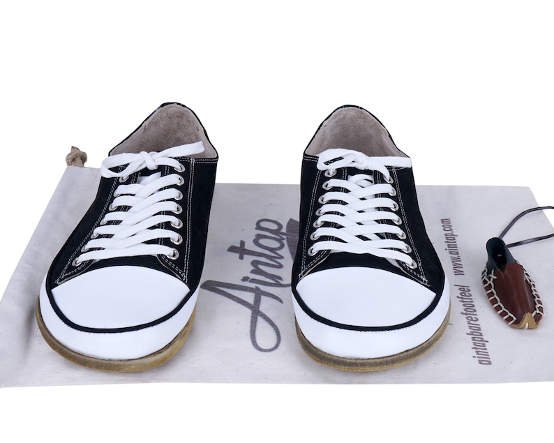 May include: Two black canvas sneakers with white laces and white soles. The shoes are sitting on a beige canvas bag with the word 'Ainto' printed on it. The bag also has the text 'aintnoprefootfeel' and 'www.ainttop.com' printed on it.