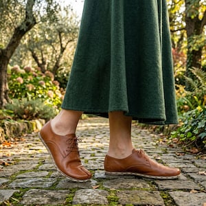 Op de afbeelding: Bruine leren schoenen met gespleten tenen en veters. De schoenen worden buiten gedragen op een stenen pad. Ze hebben een uniek ontwerp met een apart gedeelte voor de grote teen. Ze worden gecombineerd met een donkergroene rok.