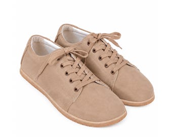 May include: A pair of low-top sneakers in a solid tan color. These shoes feature a rounded toe, matching tan laces threaded through metal eyelets, and a flat, light-colored rubber sole. The interior lining of the sneakers is white.