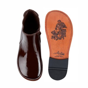 May include: A dark brown patent leather ankle boot with a side zipper and a tan leather sole. The sole features a black rubber heel piece and a stamped illustration of a shoemaker with the text Aintap Handmade in Gaziantep-Türkiye.