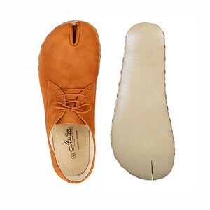Puede incluir: Una vista aérea de un par de zapatos de ante naranja con puntera dividida. Un zapato está atado con cordones y el otro es la suela. La suela es beige con un diseño de puntera dividida. El zapato con cordones tiene la marca "LizBra" impresa en la plantilla.