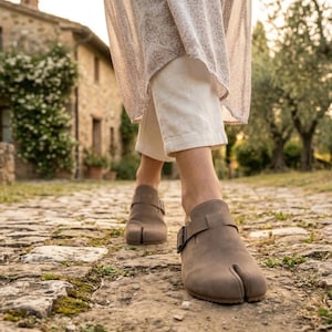May include: Taupe split-toe clogs with a buckle closure. The shoes are worn on a cobblestone path. The clogs have a strap across the top and a split between the big toe and the other toes. The person is wearing cream-colored pants.