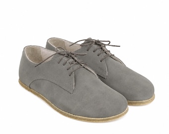 Op de afbeelding: Een paar grijze veterschoenen met een gladde matte afwerking, ronde neuzen en dunne bijpassende grijze veters. De schoenen hebben een laag profiel met een lichtbruine, platte zool die iets uitsteekt voorbij de rand van het bovenwerk.