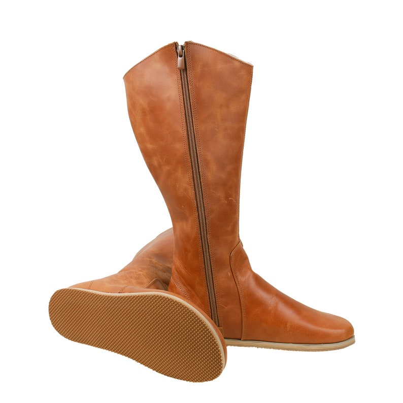 May include: A pair of knee-high, tan leather boots. One boot is upright, with a full-length zip, while the other lies horizontally, showing the textured sole. The boots have a classic, minimalist design.