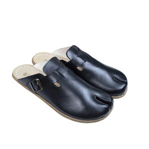 May include: A pair of black leather clogs with a split toe design. The clogs have a buckle closure and a light-colored insole. The soles are a light brown color.