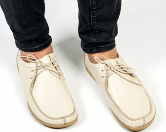 May include: A pair of off-white leather lace-up shoes with a rounded toe and a stitched seam around the edge. The shoes are worn by a person wearing dark blue jeans.