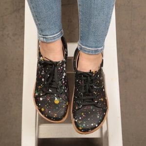 Op de afbeelding: Zwarte canvas sneakers met een kleurrijk verfspatten ontwerp. De schoenen hebben veters en een bruine rubberen zool.