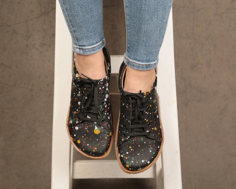 Op de afbeelding: Zwarte canvas sneakers met een kleurrijk verfspatten ontwerp. De schoenen hebben veters en een bruine rubberen zool.