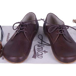K&ouml;nnte beinhalten: Ein Paar braune Lederschn&uuml;rschuhe mit abgerundeter Spitze. Die Schuhe befinden sich auf einer wei&szlig;en Oberfl&auml;che mit dem aufgedruckten Text "Hint".