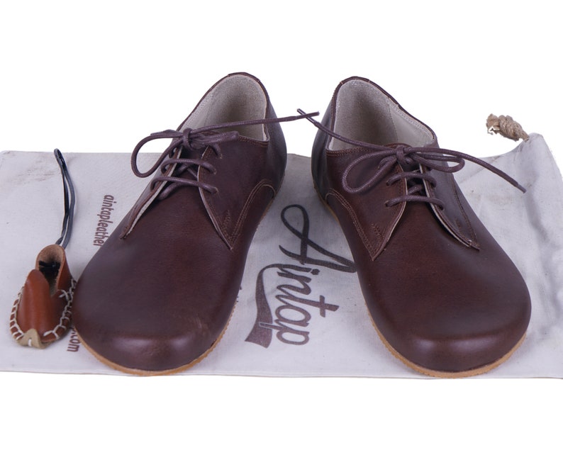 K&ouml;nnte beinhalten: Ein Paar braune Lederschn&uuml;rschuhe mit abgerundeter Spitze. Die Schuhe befinden sich auf einer wei&szlig;en Oberfl&auml;che mit dem aufgedruckten Text "Hint".