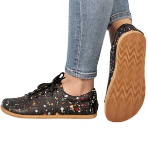 Op de afbeelding: Zwarte canvas sneakers met een kleurrijk verfspatten ontwerp. De schoenen hebben een bruine rubberen zool en worden gedragen door iemand die blauwe jeans draagt.