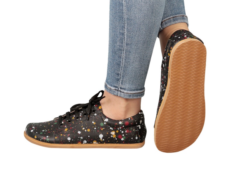 Op de afbeelding: Zwarte canvas sneakers met een kleurrijk verfspatten ontwerp. De schoenen hebben een bruine rubberen zool en worden gedragen door iemand die blauwe jeans draagt.