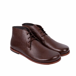 May include: A pair of brown leather chukka boots with a textured finish. The boots have a lace-up design and a rounded toe. The soles are a lighter shade of brown. The boots are displayed on a white background.