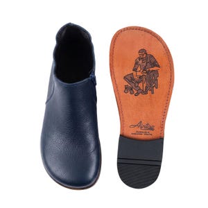 May include: A navy blue ankle boot with a textured leather upper and a side zipper. Beside it, the tan leather sole features a black illustration of a shoemaker at work, the brand name Aintap, and the text Handmade in Gaziantep-Türkiye.