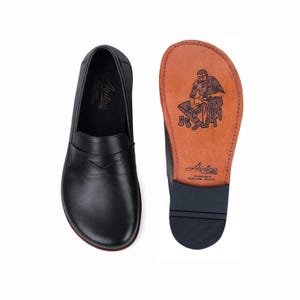 May include: Black leather loafers, one showing the sole with a craftsman illustration and the brand name, the other showing the top view. The soles are dark grey, and the inner sole is brown. The shoes are a classic design.