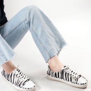 May include: A pair of black and white zebra print low-top sneakers with white laces. The sneakers are worn by a person wearing light blue jeans.