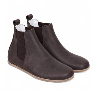May include: A pair of dark brown ankle boots featuring a smooth matte finish, elastic side panels in a matching dark brown tone, a pull tab at the heel, and a flat, light-coloured sole. The interior lining of the boots is white.