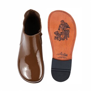 May include: A glossy brown ankle boot with a side zipper and a matching leather sole. The sole features a black rubber heel cap and a stamped illustration of a shoemaker at work, along with the text Aintap Handmade in Gaziantep-Türkiye.