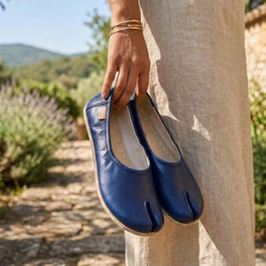May include: A pair of blue leather split-toe ballet flats. The shoes have a rounded toe and a split design for the big toe. The soles are a light brown color. The shoes are being held up by a person.