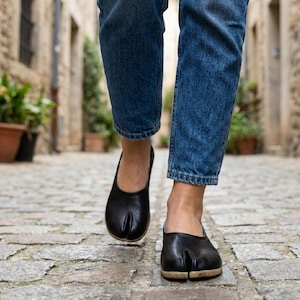 May include: Black split-toe shoes with a low heel. The shoes are worn with blue denim jeans. The shoes have a unique design with a split between the big toe and the other toes. The shoes are made of a smooth, dark material.