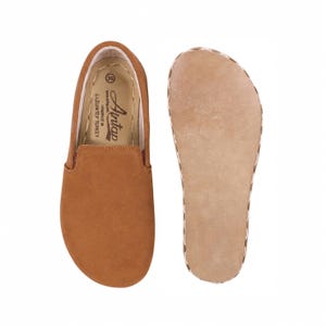 May include: A pair of tan suede slip-on shoes with a rounded toe. The interior insole features the text Antap, size 39, and handmade in Gaziantep Turkey. The second shoe is shown from the underside, revealing a flat, light tan leather sole.