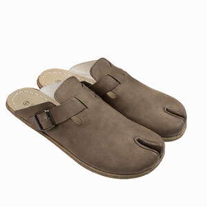 May include: A pair of brown leather clogs with a split-toe design. The clogs have a strap with a metal buckle for adjustment and a light-colored insole. The soles are a lighter brown color.