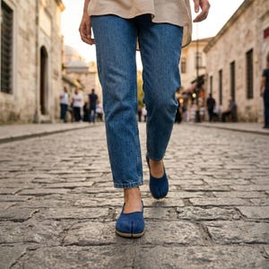 May include: A person walking on a cobblestone street wearing blue denim jeans and navy blue split-toe shoes. The shoes have a low heel and a tan sole. The background shows a European-style street with stone buildings.