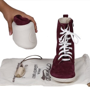 May include: Burgundy suede ankle boots with white laces and a zipper on the side. The boots are on a white canvas bag with the word "Tiwop" written on it. The bag is on a white surface.