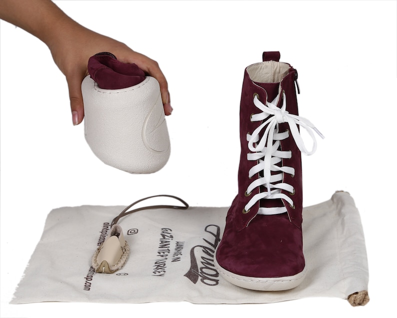 May include: Burgundy suede ankle boots with white laces and a zipper on the side. The boots are on a white canvas bag with the word "Tiwop" written on it. The bag is on a white surface.
