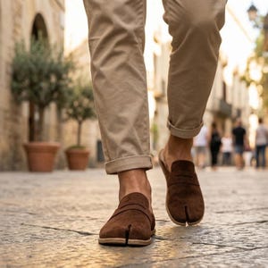 May include: Brown suede split-toe slip-on shoes with a leather strap detail. The shoes have a natural sole and are being worn with tan trousers. The image is taken outdoors on a cobbled street.