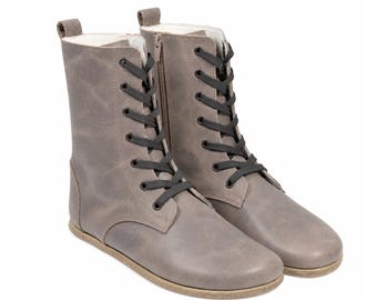 May include: A pair of mid-calf boots in a matte grey finish featuring black laces, a side zip closure, a pull tab at the heel, and a flat, light-coloured sole. The boots have a rounded toe and a smooth, uniform texture throughout the exterior.