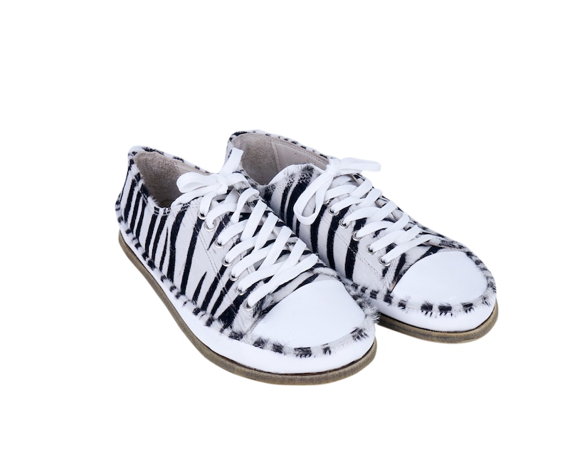 May include: A pair of white canvas sneakers with black zebra print. The shoes have white laces and a white rubber toe cap.