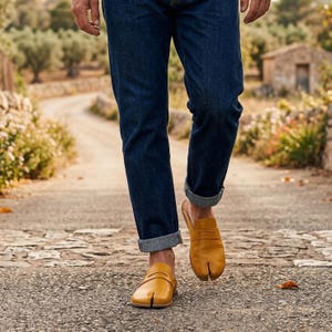 May include: A person wearing blue denim jeans with rolled-up cuffs and mustard-yellow split-toe leather shoes. The shoes have a slip-on design with a separate section for the big toe. The person is walking on a stone path.