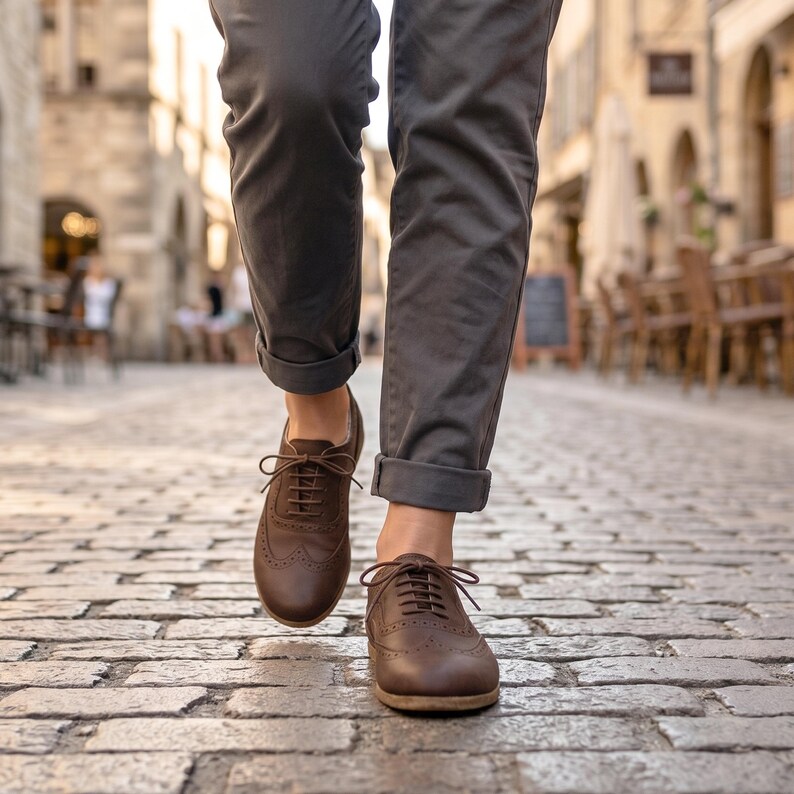 May include: A pair of dark brown leather oxford shoes with wingtip brogue detailing and matching brown laces, worn with charcoal gray cuffed trousers. The shoes feature a rounded toe and a low heel, showcasing a classic lace-up design.