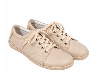 May include: A pair of low-top sneakers in a solid beige color. These shoes feature a smooth exterior, matching beige laces threaded through metal eyelets, a rounded toe, and a flat, tonal rubber sole. The interior lining is a light off-white color.