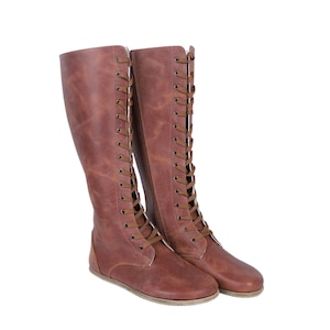 Könnte beinhalten: Ein Paar kniehohe braune Lederstiefel mit flacher Sohle. Die Stiefel haben ein Schnürdesign mit braunen Schnürsenkeln und Ösen. Das Leder hat einen Used-Look, und die Stiefel sind vor weißem Hintergrund platziert.