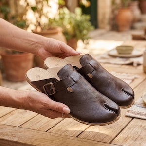 May include: A pair of brown leather clogs with a buckle closure. The clogs have a split toe design and a light-colored insole. The shoes are held by hands, showcasing the footwear's details and craftsmanship.