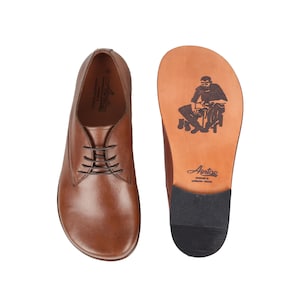 Könnte beinhalten: Braune Lederschuhe mit Schnürung. Die Sohle eines Schuhs zeigt eine gedruckte Illustration einer Person und den Markennamen "Antap". Die Schuhe sind für Komfort und Stil konzipiert, passend für verschiedene Anlässe.