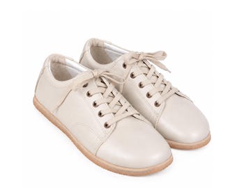 May include: A pair of low-top sneakers in a solid off-white color. These shoes feature a smooth upper, matching off-white laces threaded through metal eyelets, a rounded toe, and a flat, light tan rubber sole with a textured edge for traction.