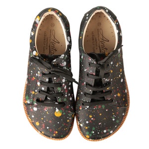 Op de afbeelding: Een paar zwarte canvas sneakers met een kleurrijk splatterontwerp. De schoenen hebben zwarte veters en een bruine rubberen zool. De tekst "Ainto" staat gedrukt aan de binnenkant van de schoenen.