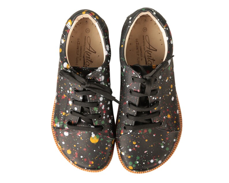Op de afbeelding: Een paar zwarte canvas sneakers met een kleurrijk splatterontwerp. De schoenen hebben zwarte veters en een bruine rubberen zool. De tekst "Ainto" staat gedrukt aan de binnenkant van de schoenen.