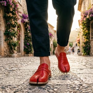 May include: Red leather split-toe slip-on shoes with a wooden sole. The shoes are being worn on a cobbled street. The person is wearing dark blue trousers. The background shows a European style street with buildings and flowers.