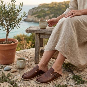 May include: Brown leather slip-on clogs with a toe separation design. The clogs have a buckle detail and are worn by a person sitting outdoors. A small wooden table holds a mug, and a potted olive tree is nearby.