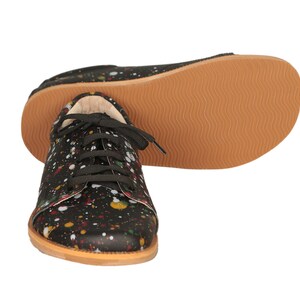 Op de afbeelding: Zwarte canvas sneakers met een kleurrijk verfspatten ontwerp. De schoenen hebben zwarte veters en een bruine rubberen zool.