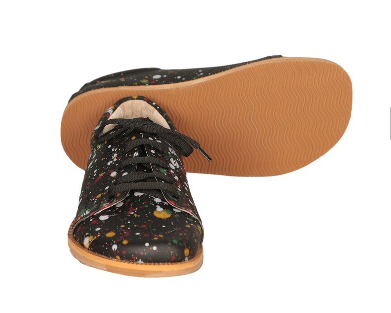 Op de afbeelding: Zwarte canvas sneakers met een kleurrijk verfspatten ontwerp. De schoenen hebben zwarte veters en een bruine rubberen zool.