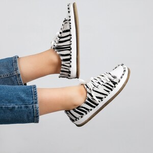 May include: A pair of white canvas sneakers with black zebra print. The sneakers have a white lace-up closure and a brown rubber sole.