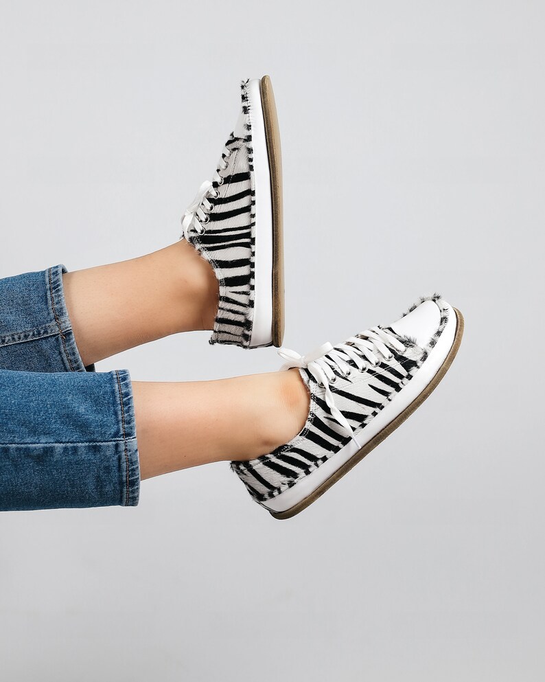 May include: A pair of white canvas sneakers with black zebra print. The sneakers have a white lace-up closure and a brown rubber sole.